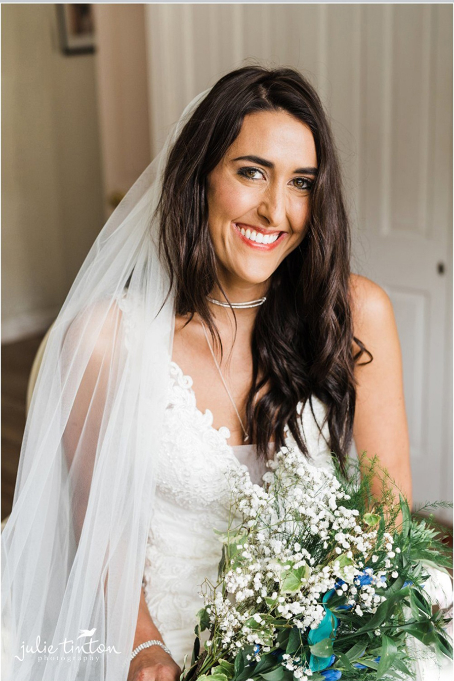 Bride smiles in hotel room with bouquet in hand and veil draped over her shoulder, her makeup is done by Jennifer Louise Studio.